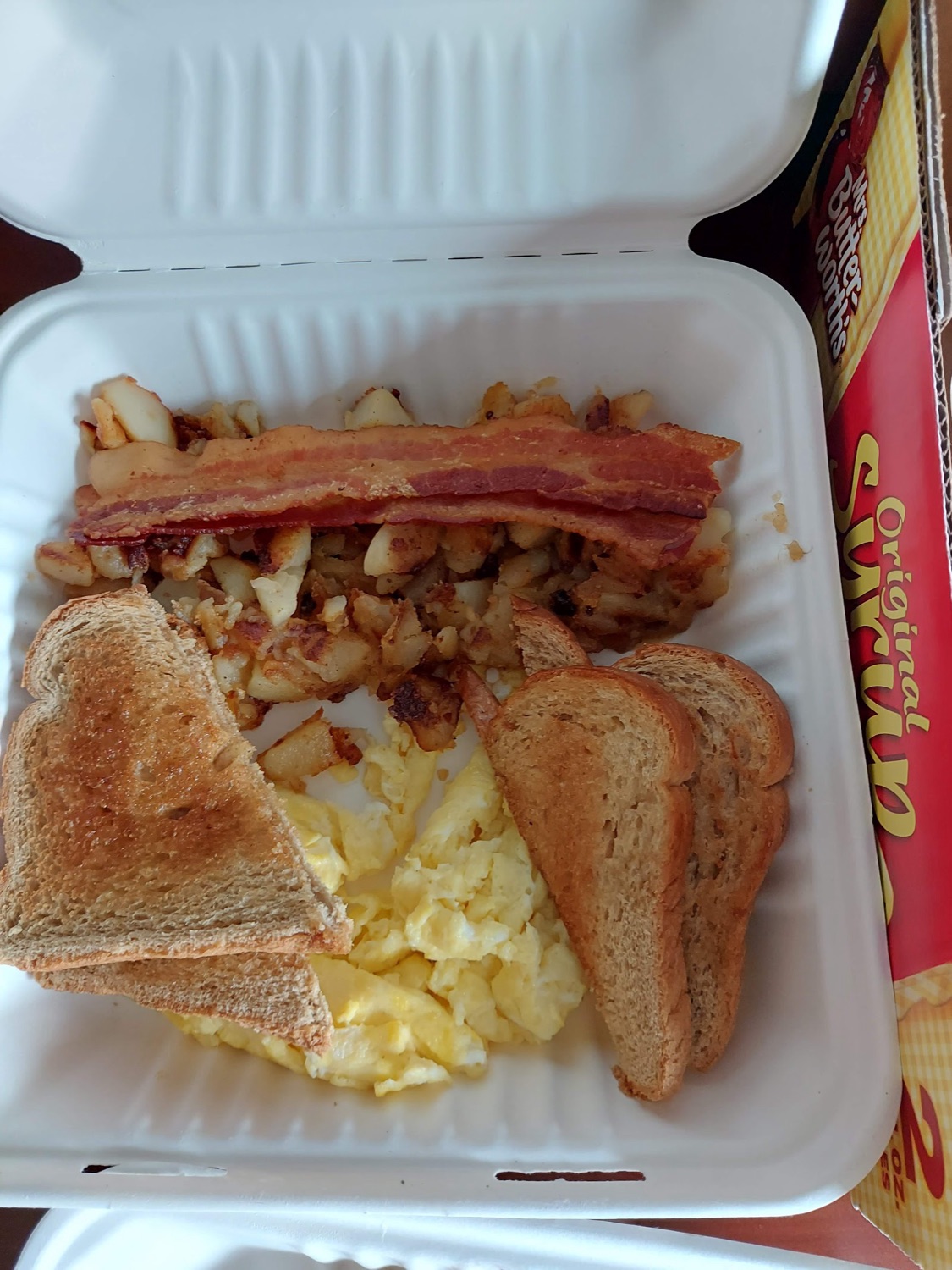 Scrambled eggs, bacon, home fries, and wheat toast in a takeout container.