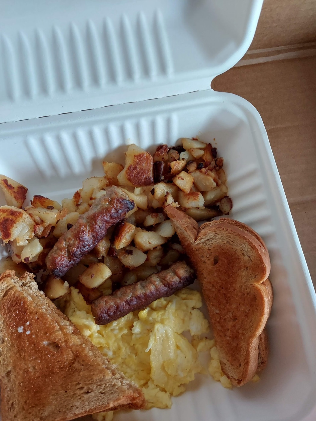 Scrambled eggs, sausage links, home fries, and toast in a takeout container.