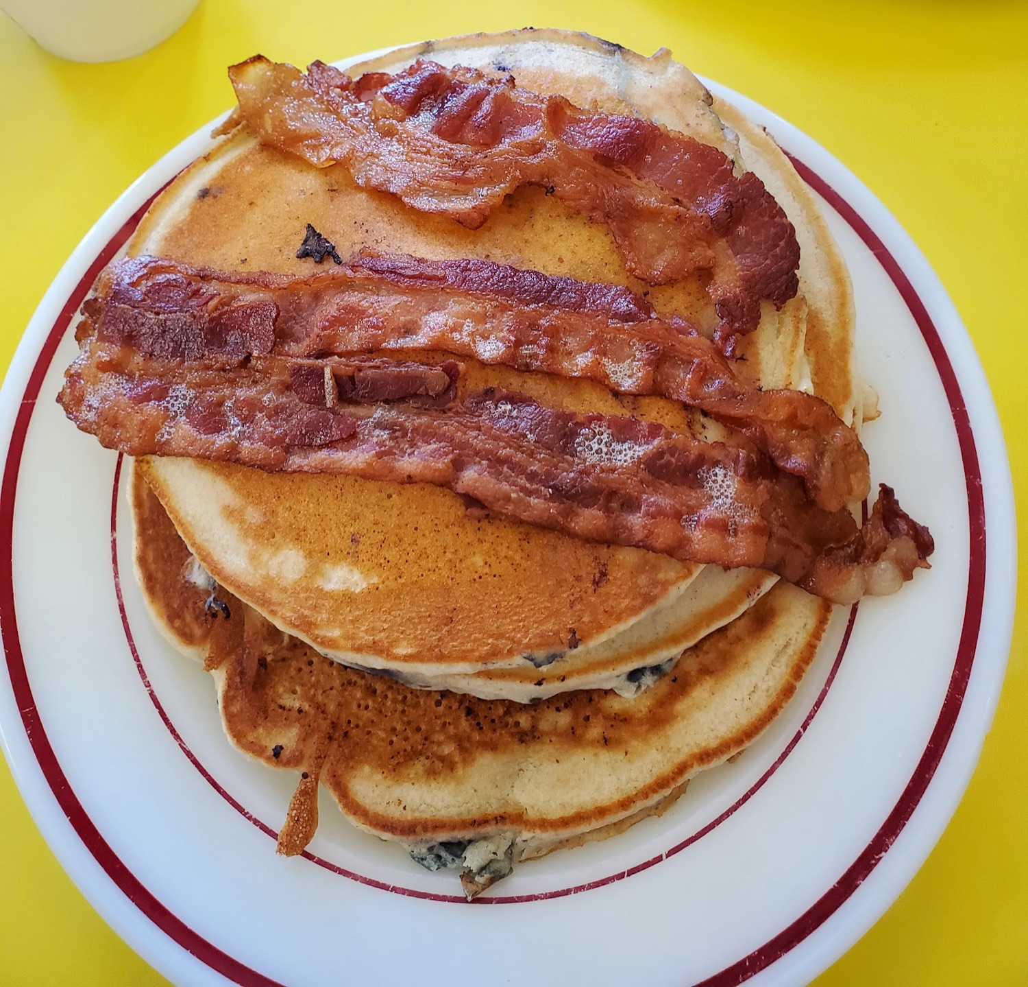 A stack of blueberry pancakes topped with strips of crispy bacon on a red-rimmed diner plate.